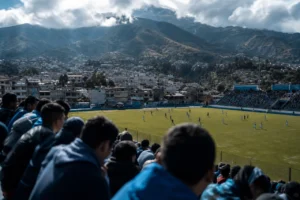 Partido de tarde en un estadio del altiplano: aficionados de espaldas en la grada y, al fondo, casas y montañas.