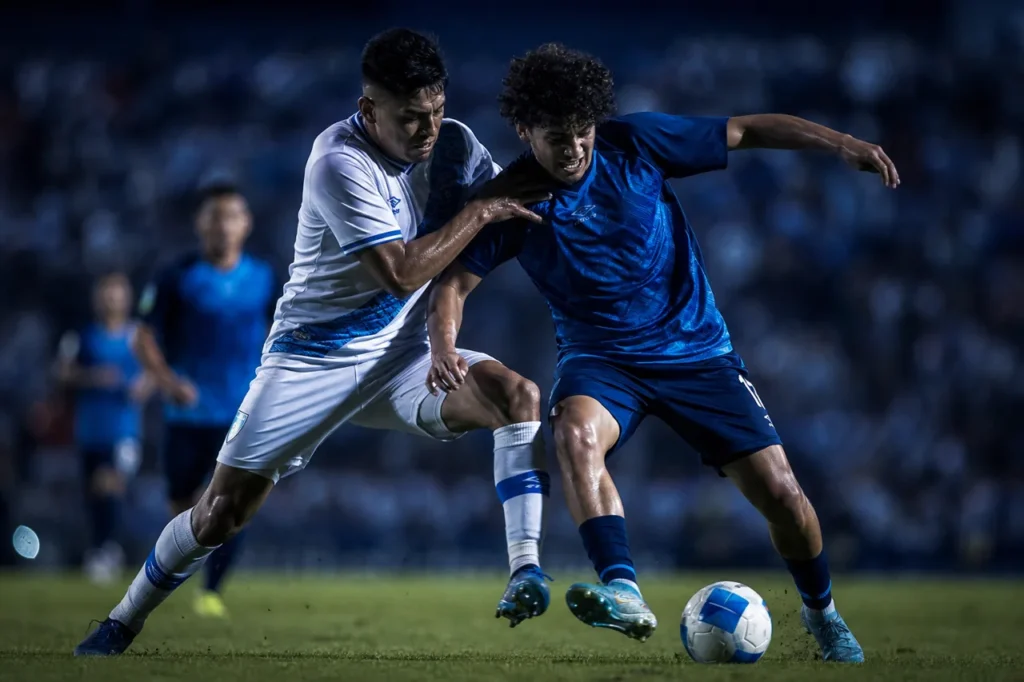 Dos futbolistas, uno de blanco y otro de azul oscuro, disputan el balón en un partido nocturno.