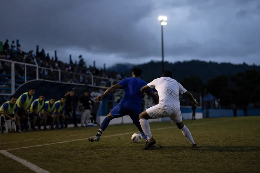 Dos futbolistas disputan un balón junto a la línea de banda en un estadio iluminado, con el banquillo desenfocado al fondo.