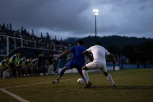Dos futbolistas disputan un balón junto a la línea de banda en un estadio iluminado, con el banquillo desenfocado al fondo.